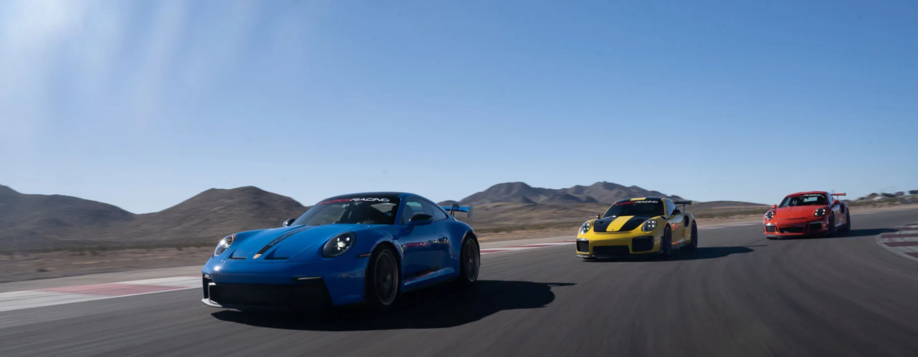 Three sports cars, a blue, yellow, and red, race on a track with mountainous desert terrain under a clear blue sky, conveying speed and excitement.